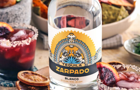 Bottle of Zarpado Blanco Tequila sitting on round wood board surrounded by cocktails, chips and salsa